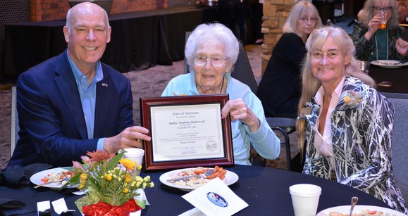 Audrey Alderwald, who turns 100 in December, has a light blue shirt and grey hair. She sits between Governor Gianforte and first lady Susan Gianforte while holding her certificate in a cherry wood frame.