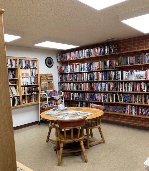 A picture of the MVH library. There are many books along the walls. One of the walls is red brick. There is a small table in the center with three chairs.
