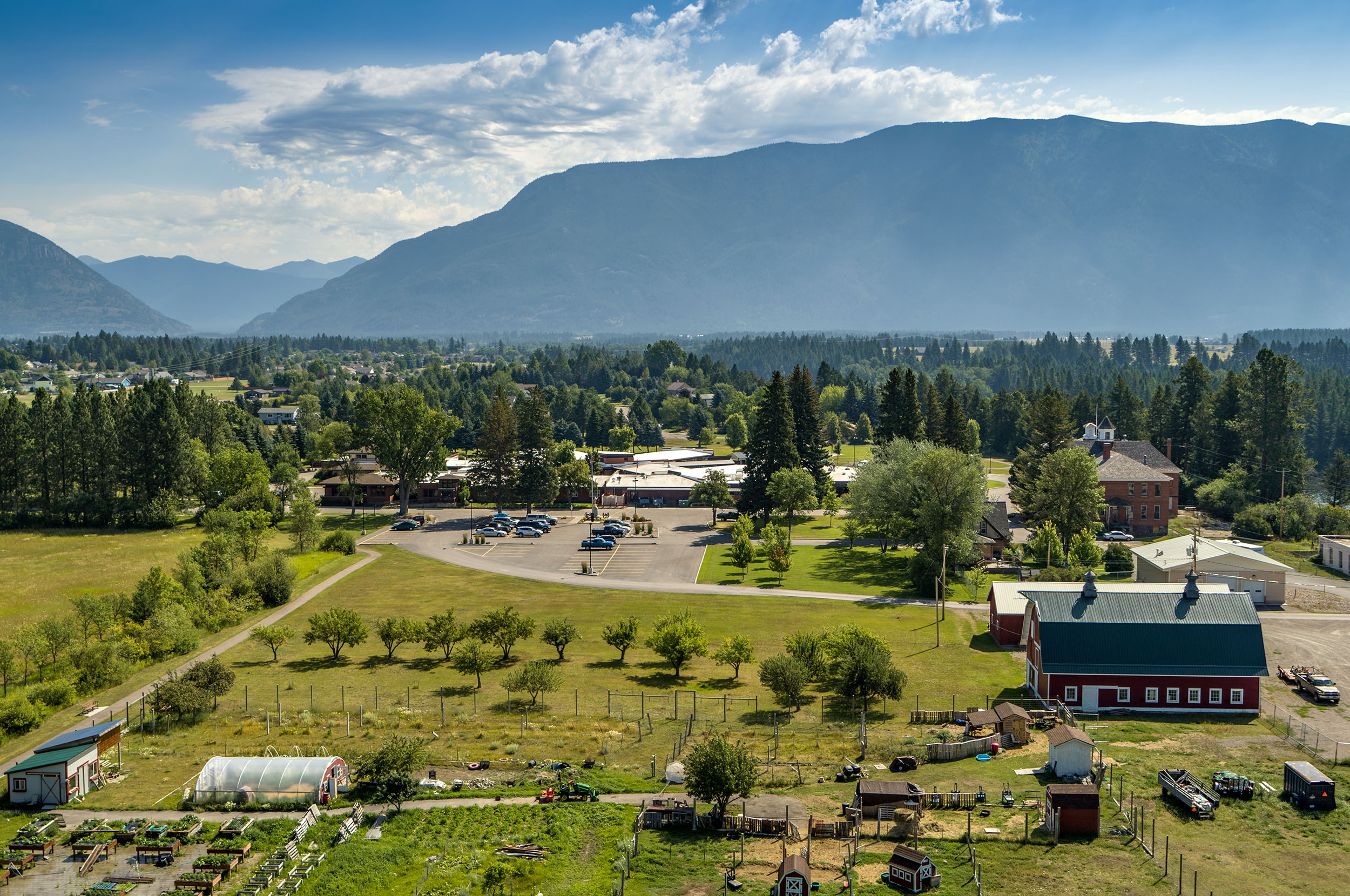 An aerial view of the grounds of the Montana Veterans home. There are large mountains in the background, as well as many trees. There is also a parking lot and several buildings.