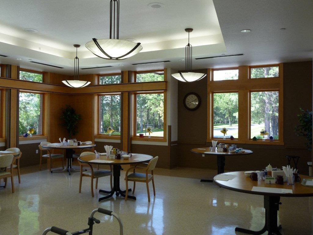 A picture of the west dining room inside the Veterans home. There are four round tables with chairs at two of them, with condiments and utensils at the center of each table. There are windows with flowers at each window, and trees outside.