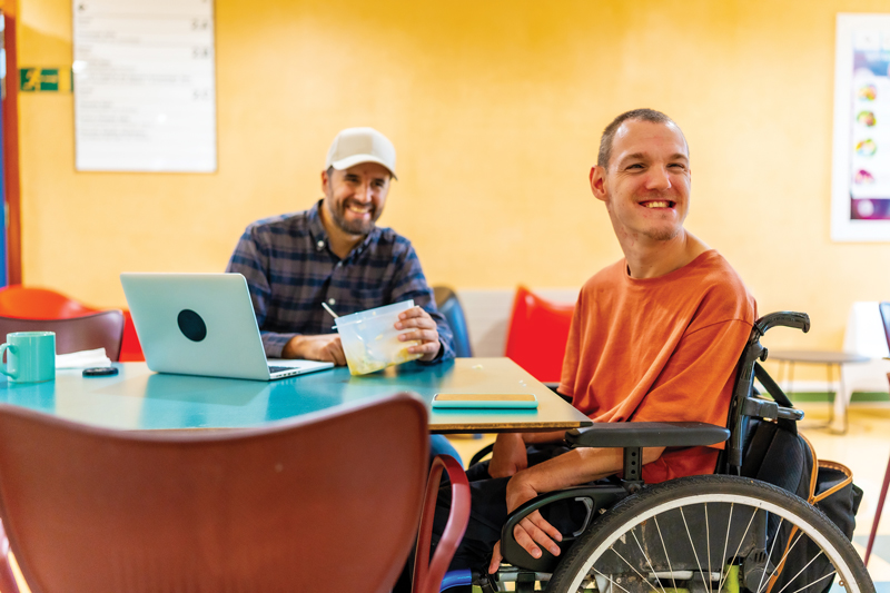 Man in a wheelchair sitting at a table next to another man. Both are smiling.